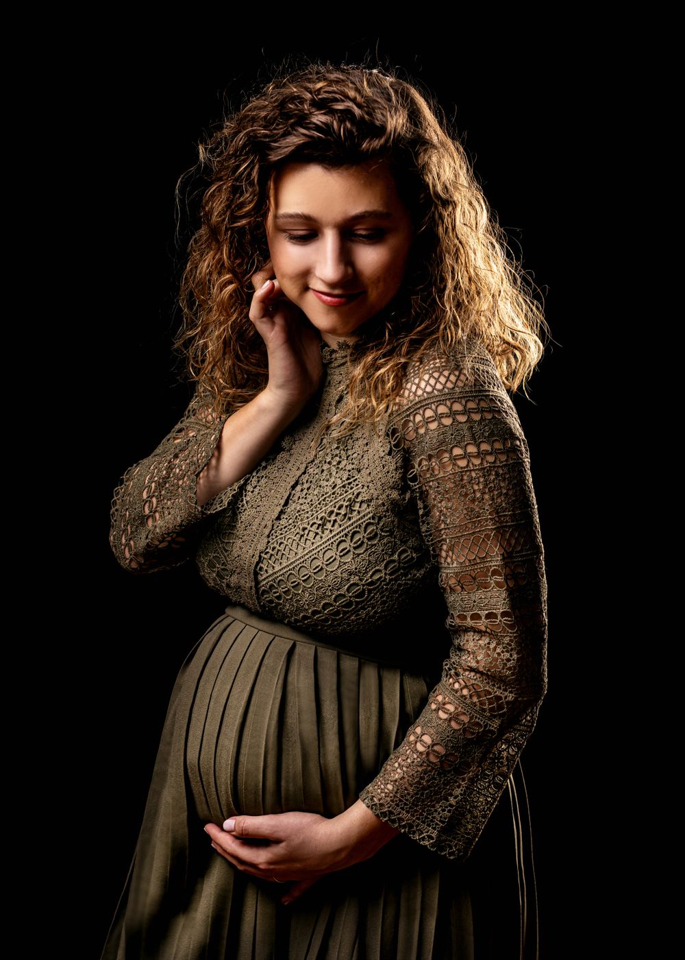 Maternity portrait of woman in brown lace dress against dark background.