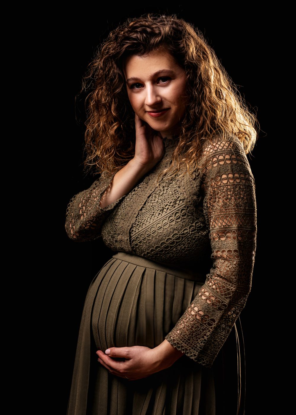 Elegant maternity photo featuring woman in brown lace dress against black backdrop.