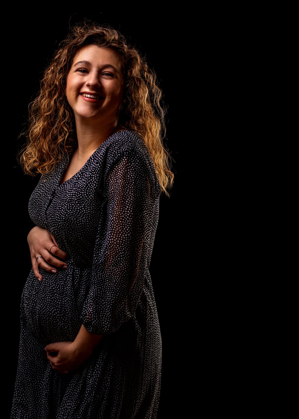Joyful maternity portrait with woman in polka dot dress against black background.
