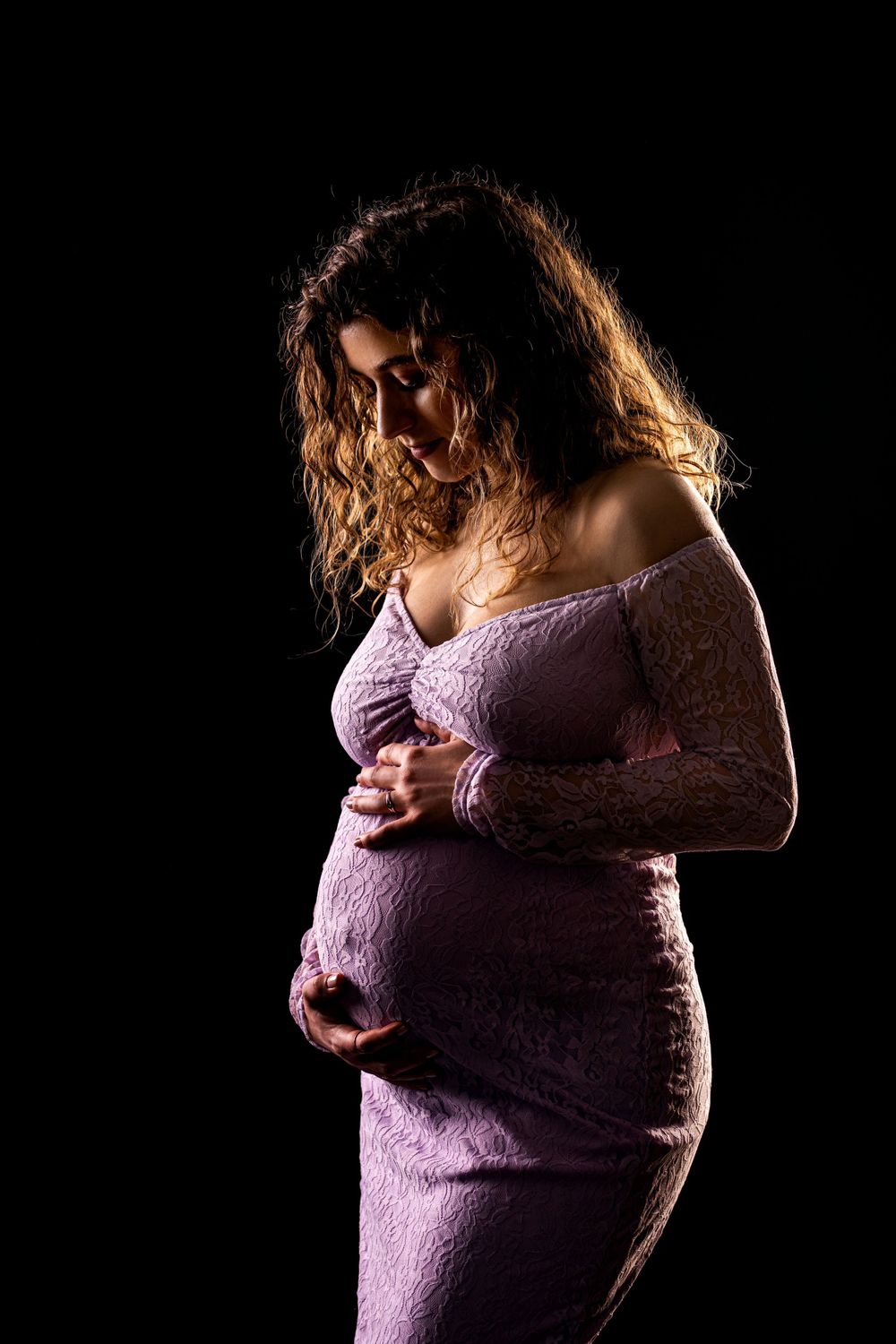 Dramatic side-lit maternity portrait of woman in purple lace dress against dark backdrop.