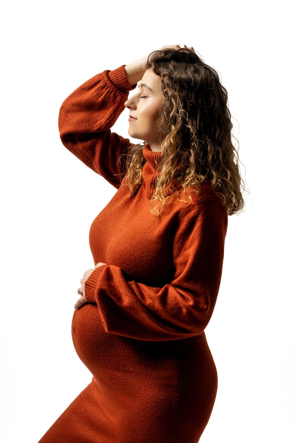 Pregnant woman in rustic orange sweater dress poses against white background with side profile.