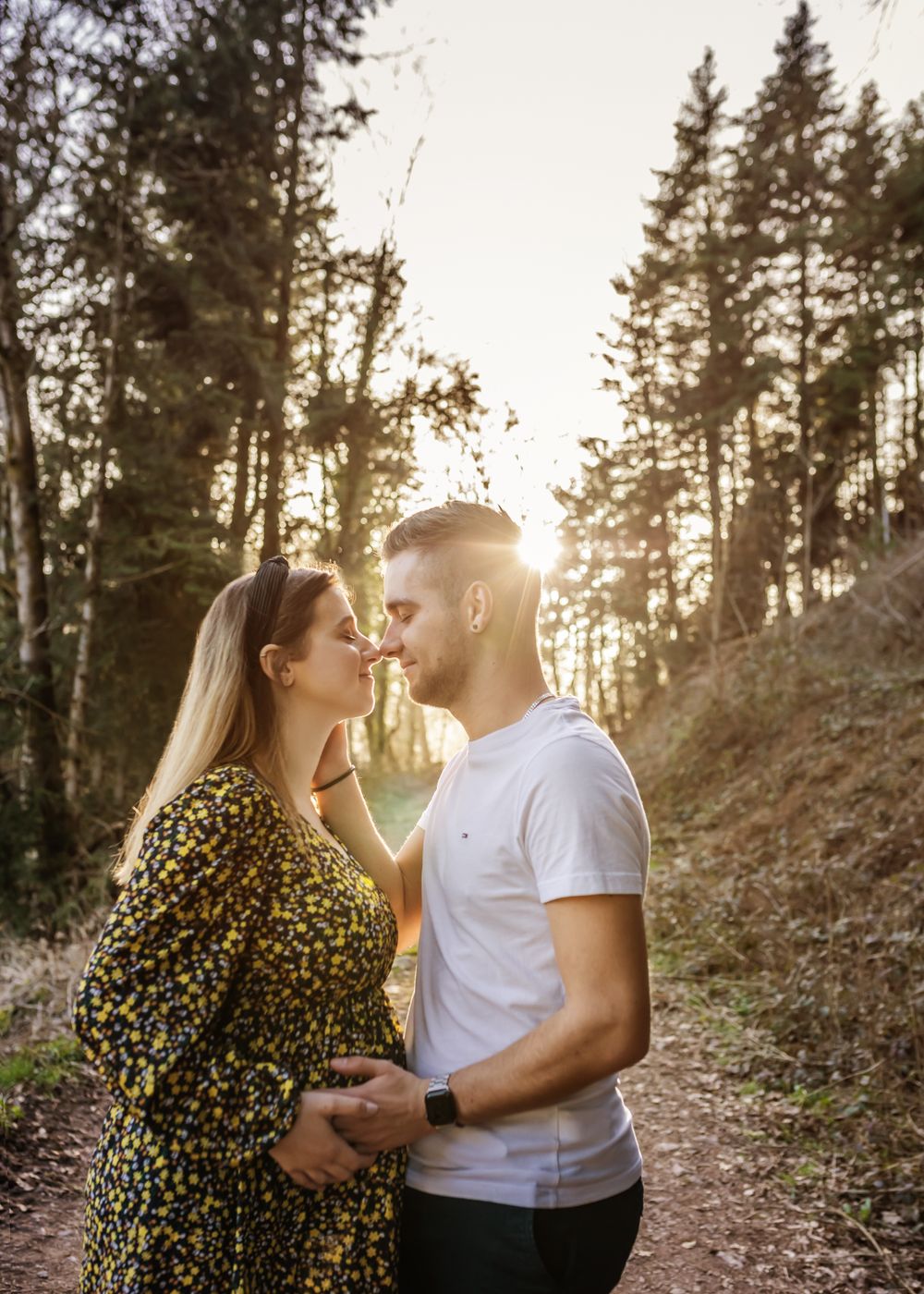 Couple embraces on forest trail during golden hour sunset with pine trees in background.