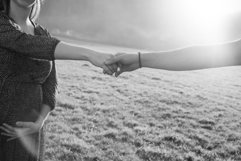 Black and white silhouette of hands reaching toward each other against sunlit meadow.