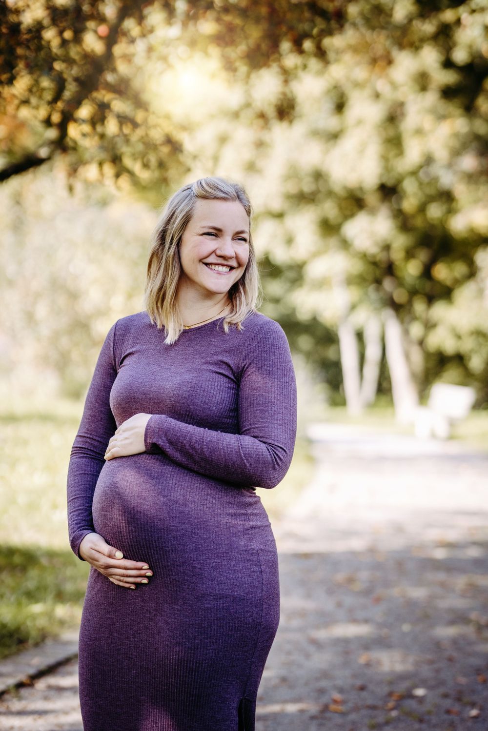 An expectant mother in a purple dress poses outdoors with sunlit trees in the background.