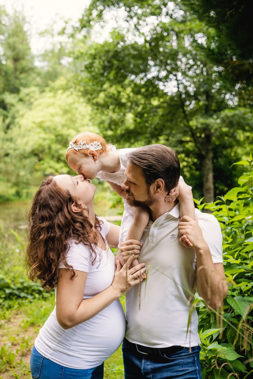 Family shares joyful moment outdoors with toddler held up high against green foliage.