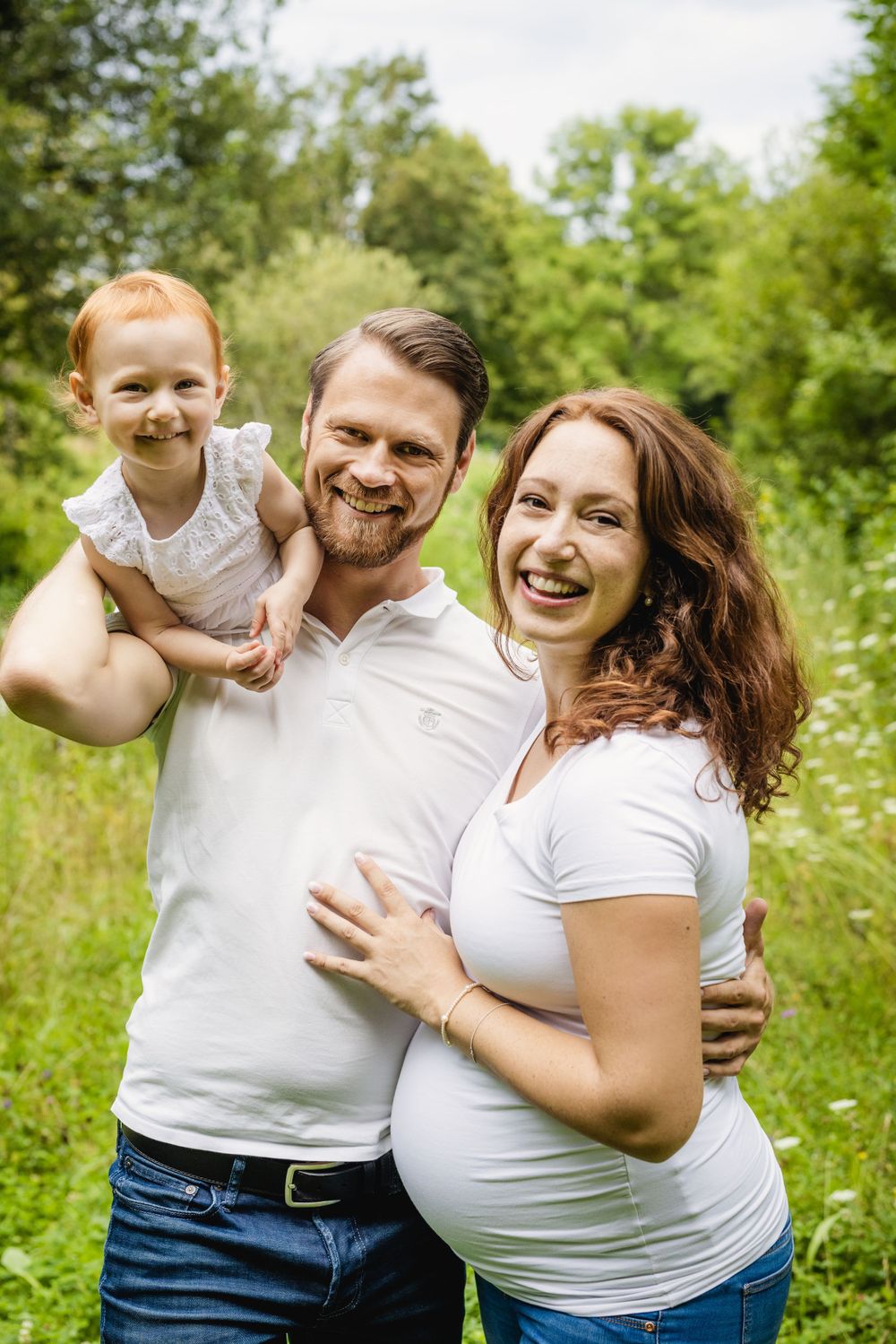 Family portrait outdoors in summer meadow showing expecting parents and young child.