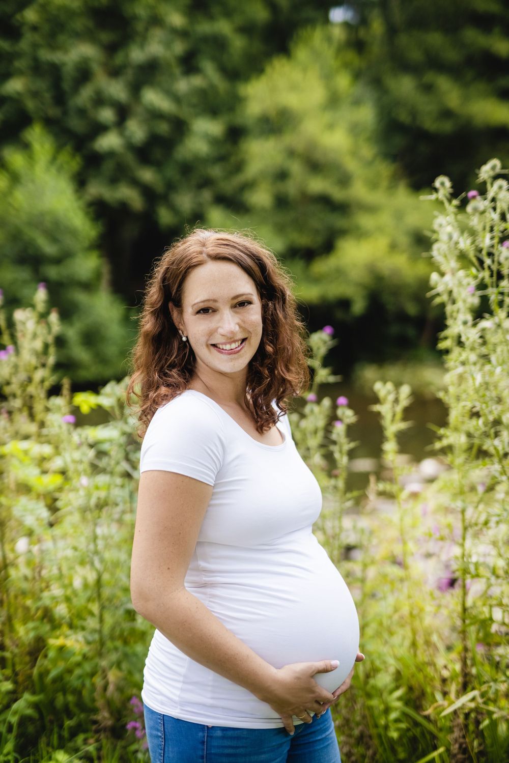 Expectant mother in white t-shirt smiles while standing in wildflower garden.