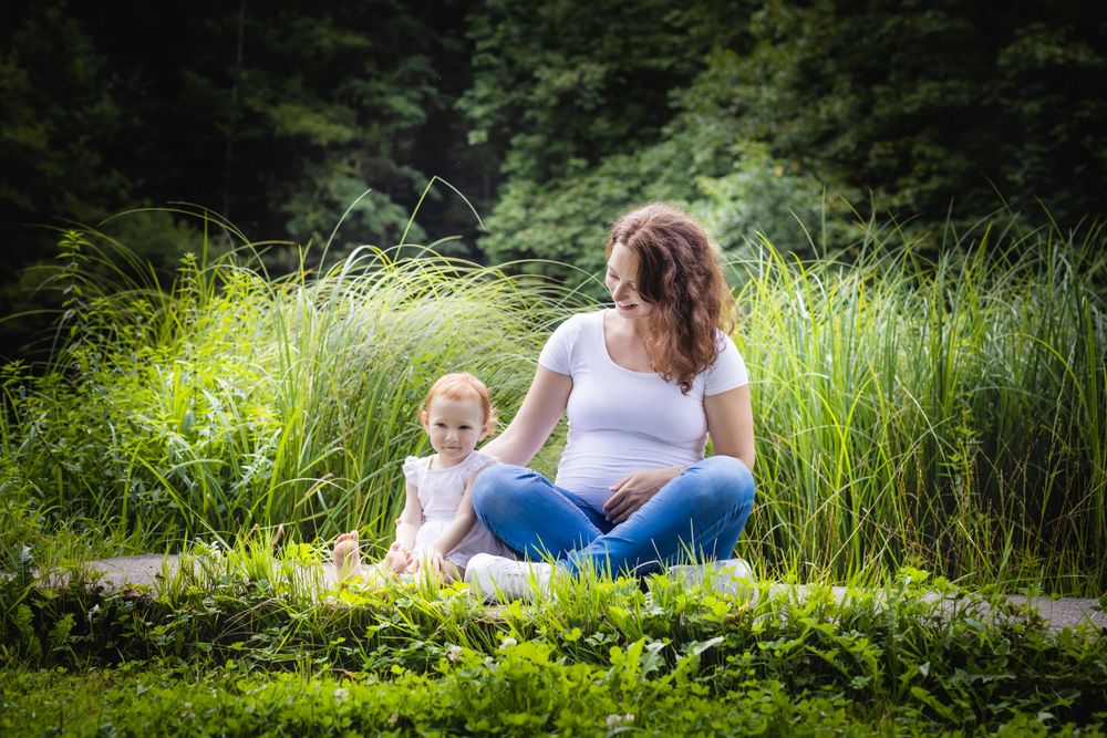 Mother and child sit together among tall grasses by pond in natural setting.
