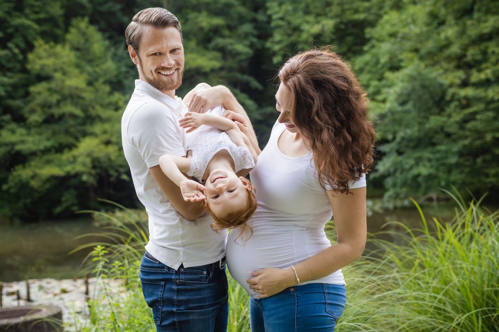 A happy family moment outdoors by a lake with parents holding and playing with their child in a natural setting.