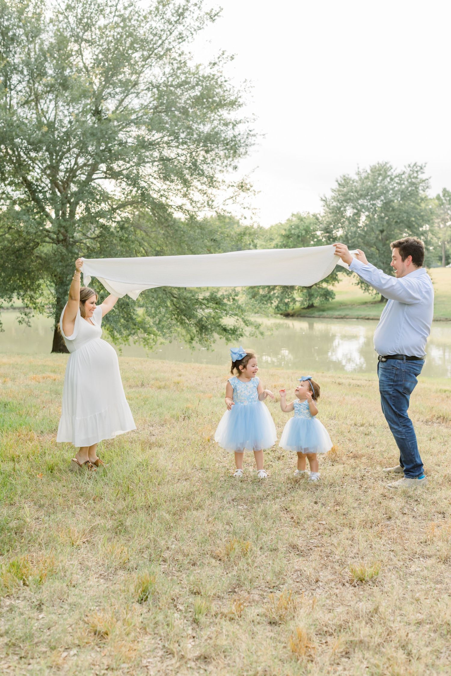 Family playing with white fabric outdoors by a pond on a sunny day.