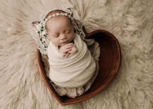 Sleeping newborn wrapped in cream swaddle lying in wooden heart bowl on fluffy white rug.