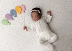 Sweet newborn in white knit outfit holding pastel colored balloons while lying on polka dot backdrop.