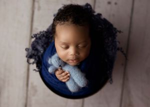 Sleeping infant in blue bassinet with curly hair holding knitted toy during newborn photoshoot.