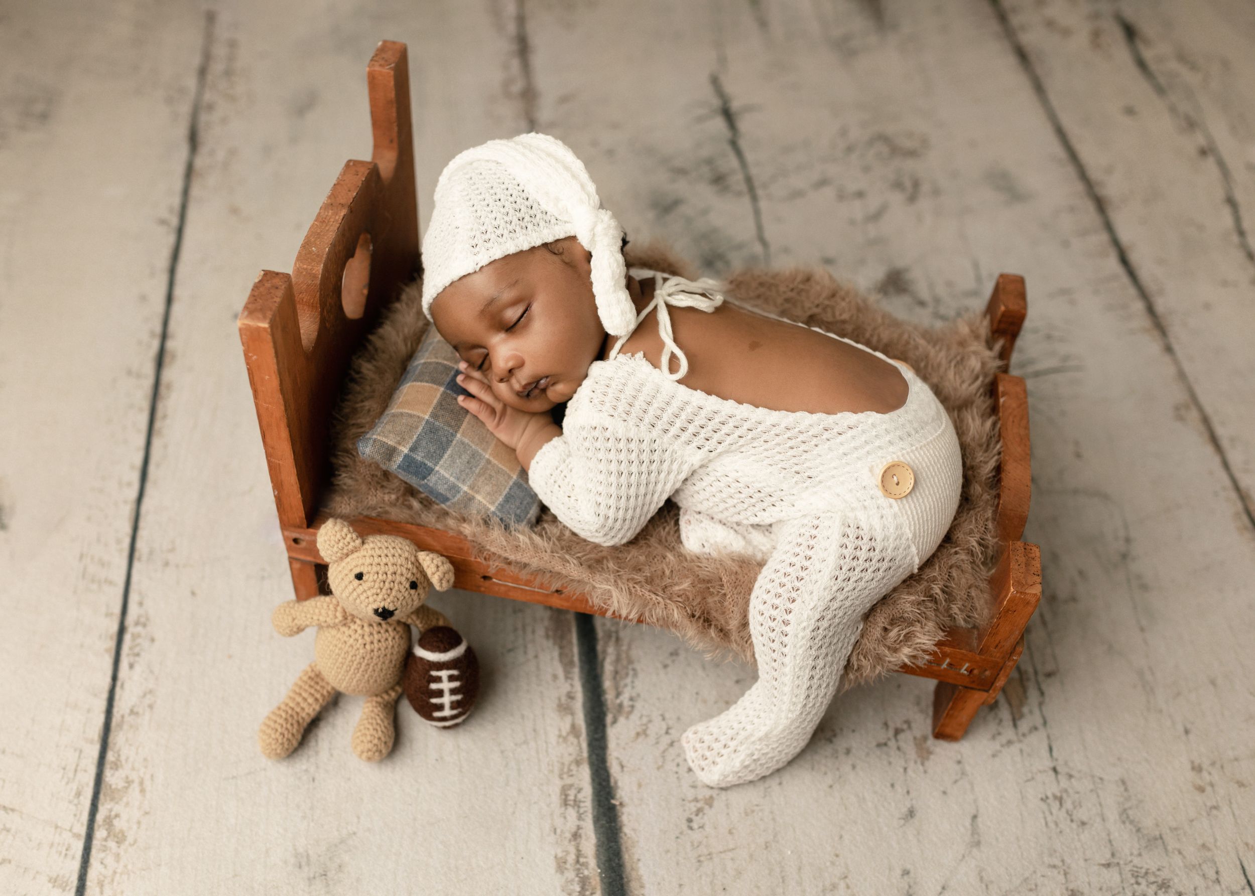 Newborn sleeping in tiny wooden bed with white knit outfit and teddy bear.