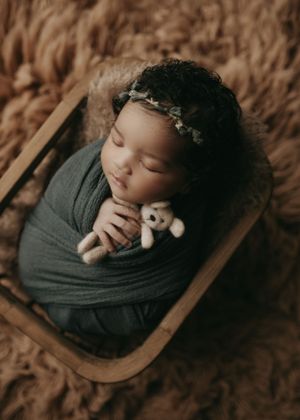Newborn baby wrapped in gray swaddle sleeping peacefully in wooden bowl with small stuffed toy.