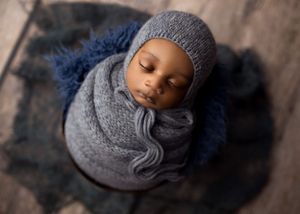 Sleeping newborn wrapped in gray knit blanket on blue furry backdrop.