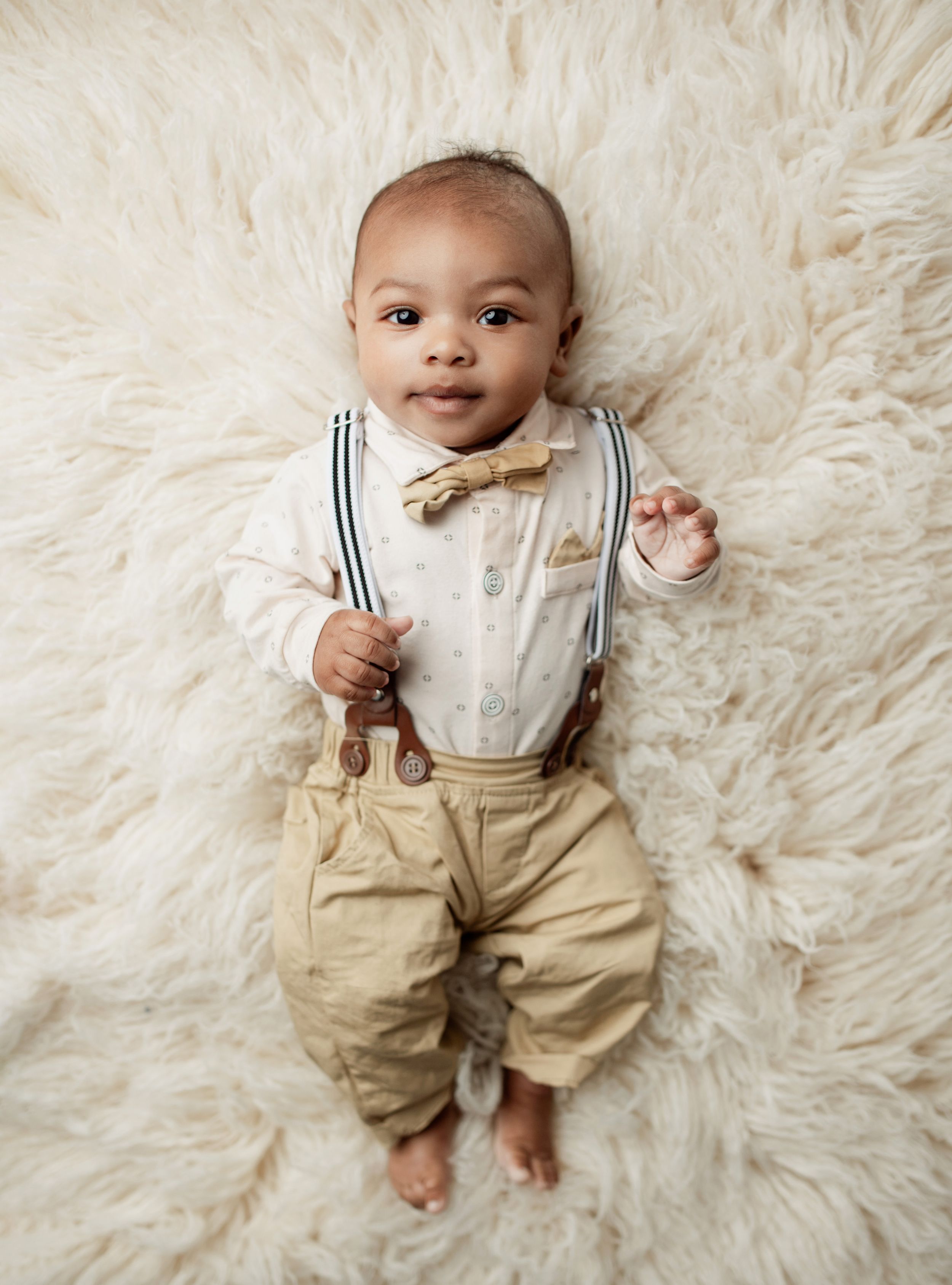 Baby wearing bow tie and suspenders outfit lying on white fluffy rug.