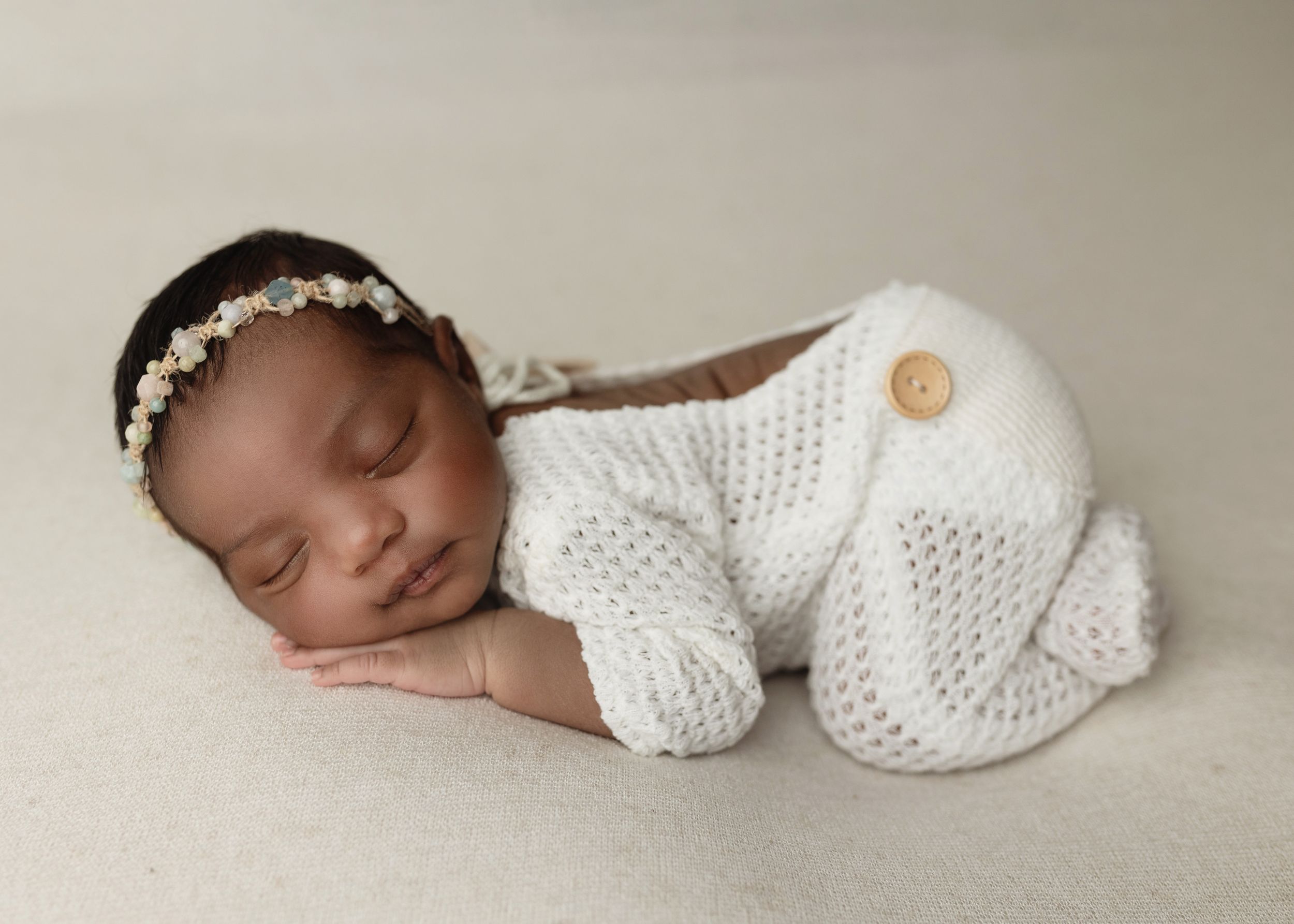Sleeping newborn in white knit outfit with floral headband on neutral backdrop.