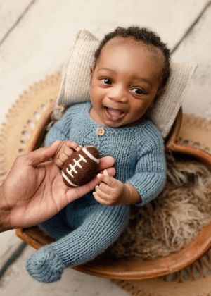 Baby in blue knit outfit holding small football toy while sitting in wooden chair.