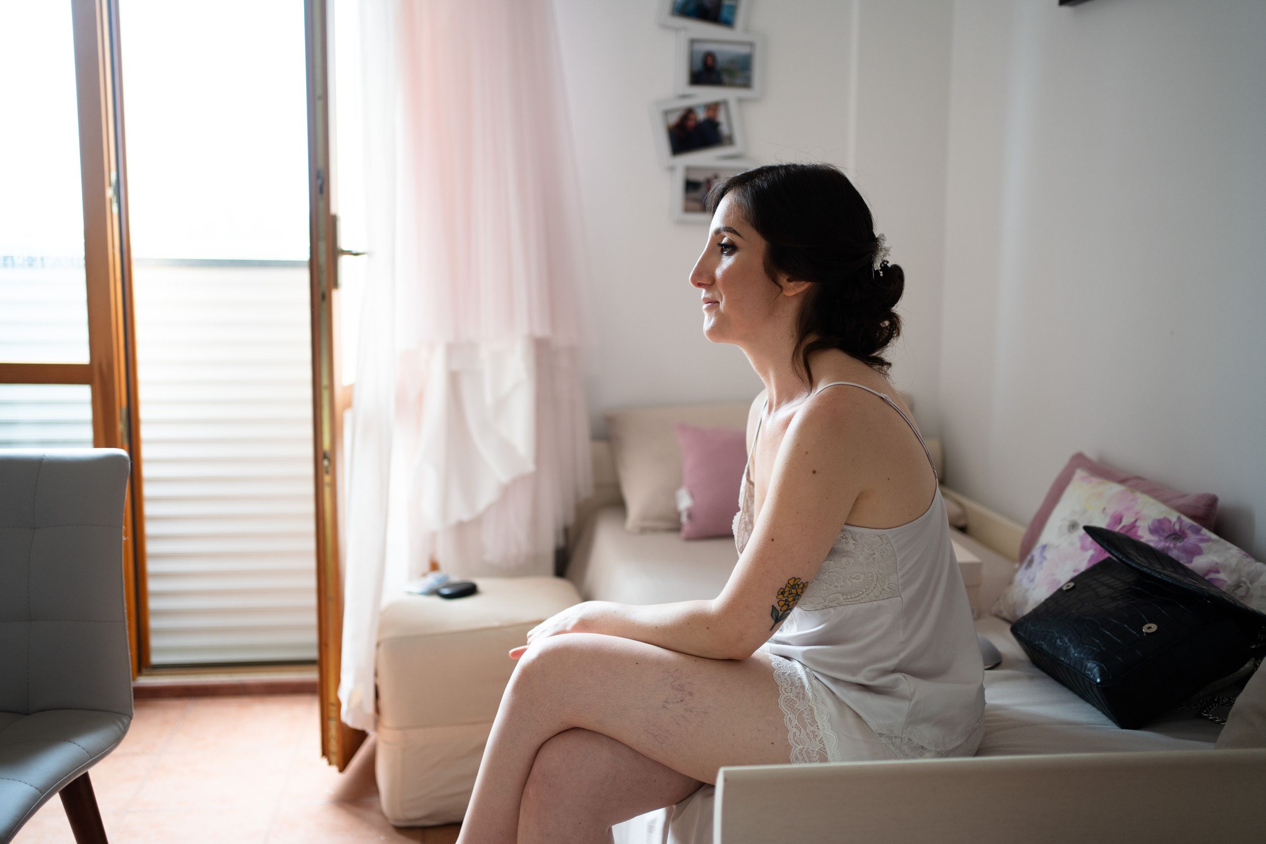 A woman in a white dress sits on a bed near a window, gazing out thoughtfully in natural daylight.