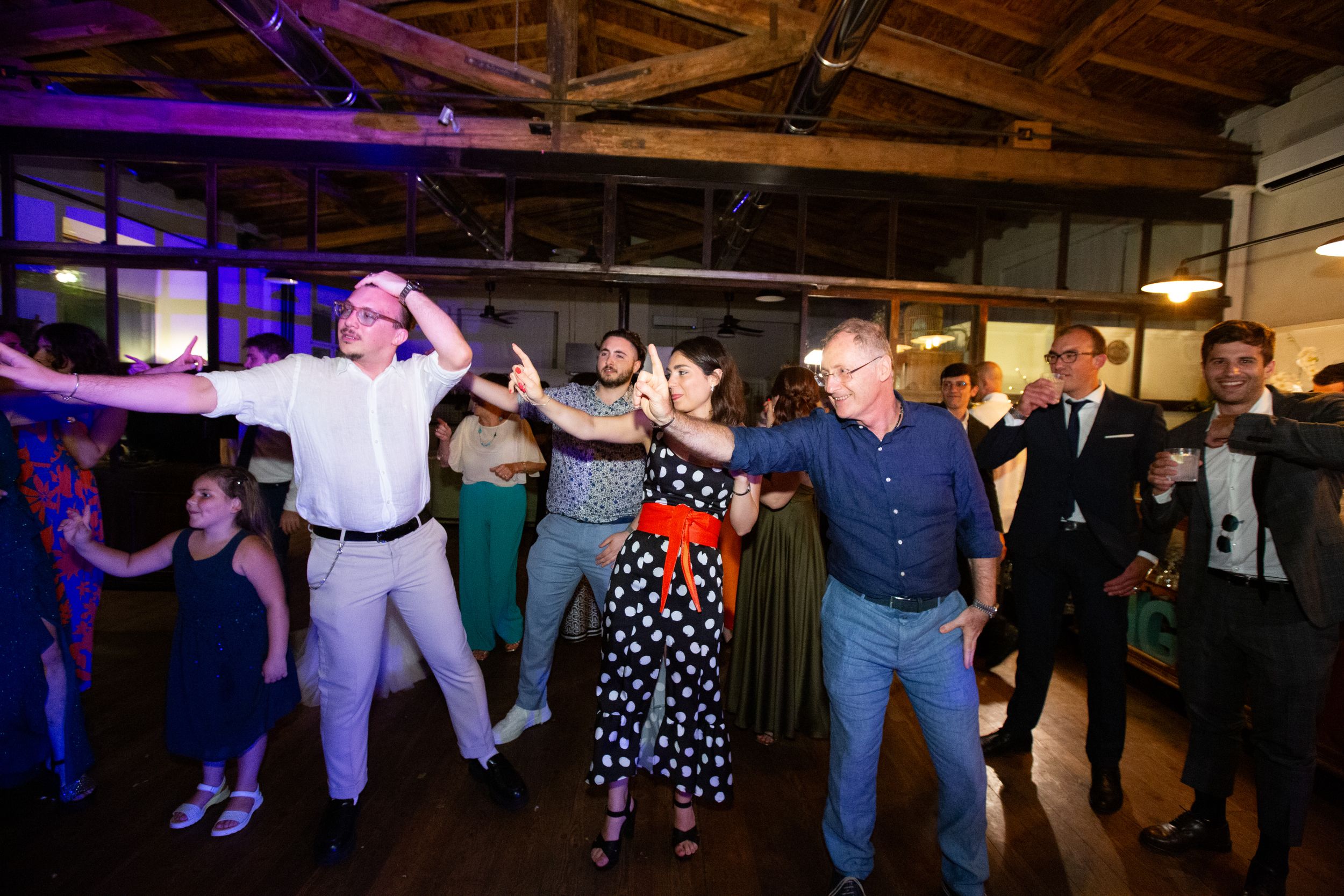Wedding party dancing together at a reception with purple lighting and exposed beams.