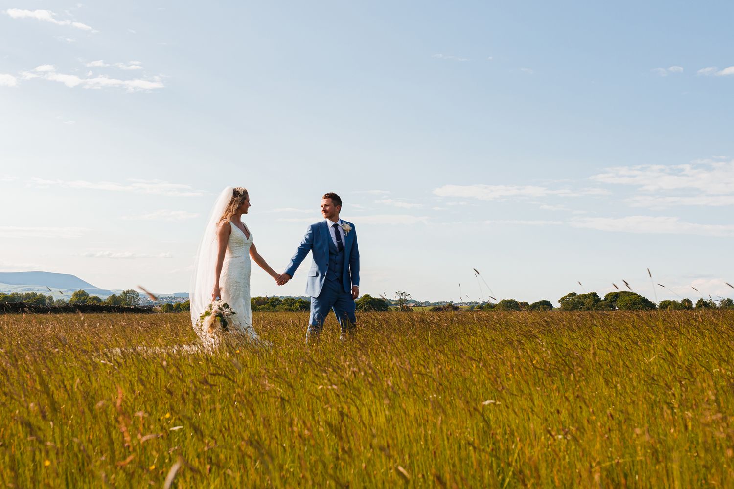 A couple walks hand in hand through a golden meadow under bright blue skies.