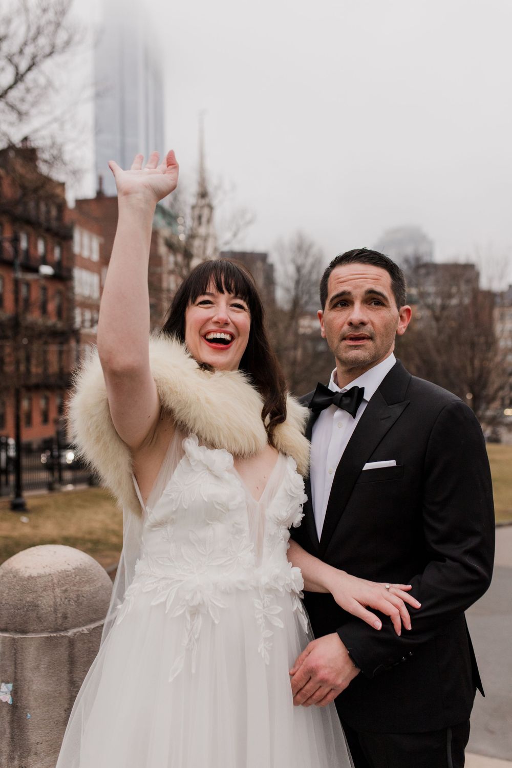 A couple dressed in wedding attire stands side by side as the bride waves at crowds cheering them on.