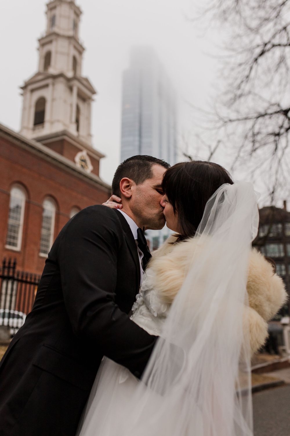 A couple dressed in wedding attire kisses in Boston Common surrounded by views of tall buildings covered in fog.