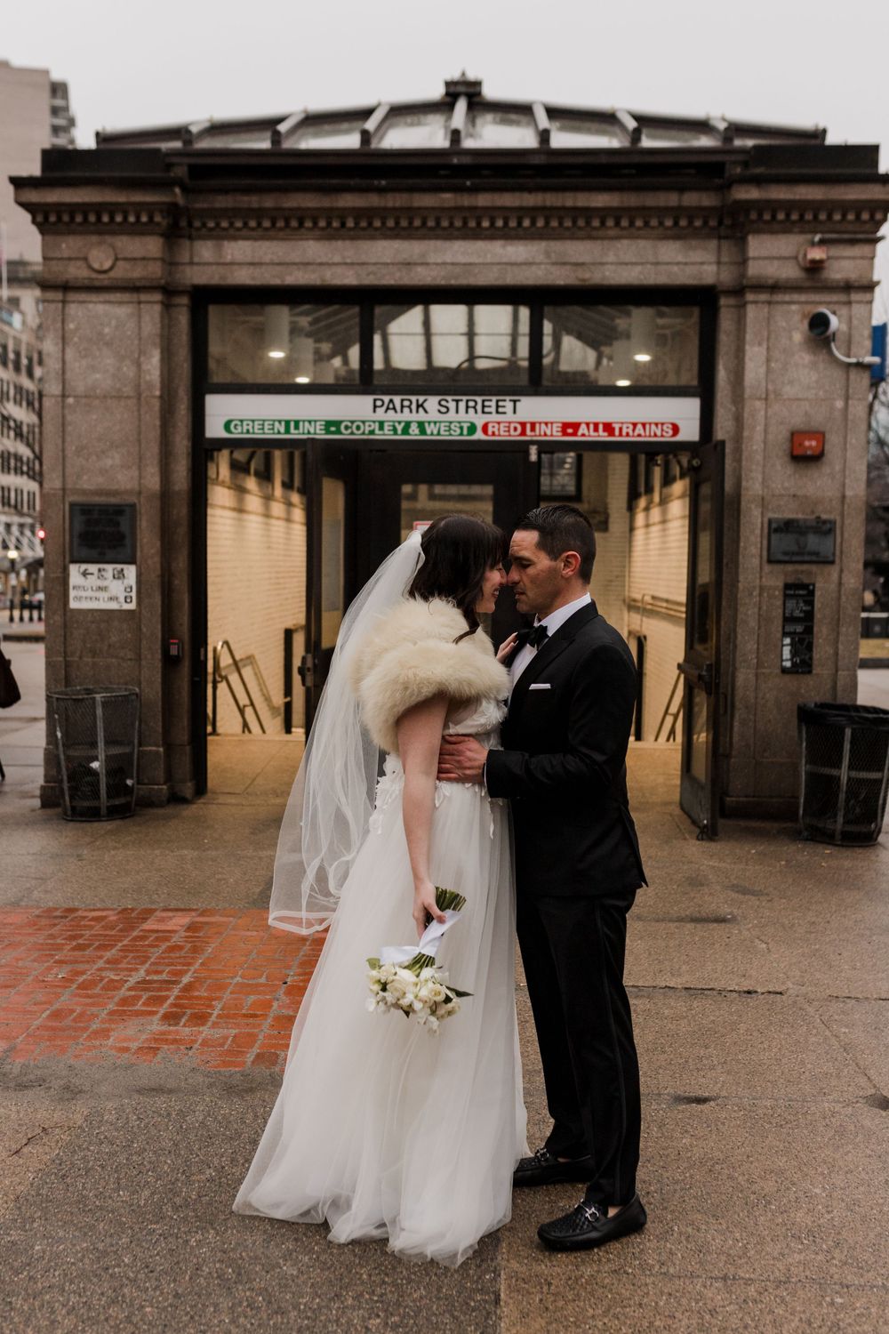 A couple dressed in wedding attire kisses in front of a Boston MBTA station.