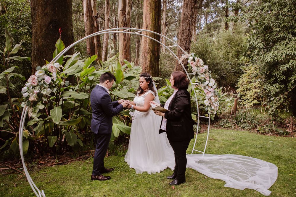 A wedding ceremony takes place under a circular floral arch in a lush forest setting.