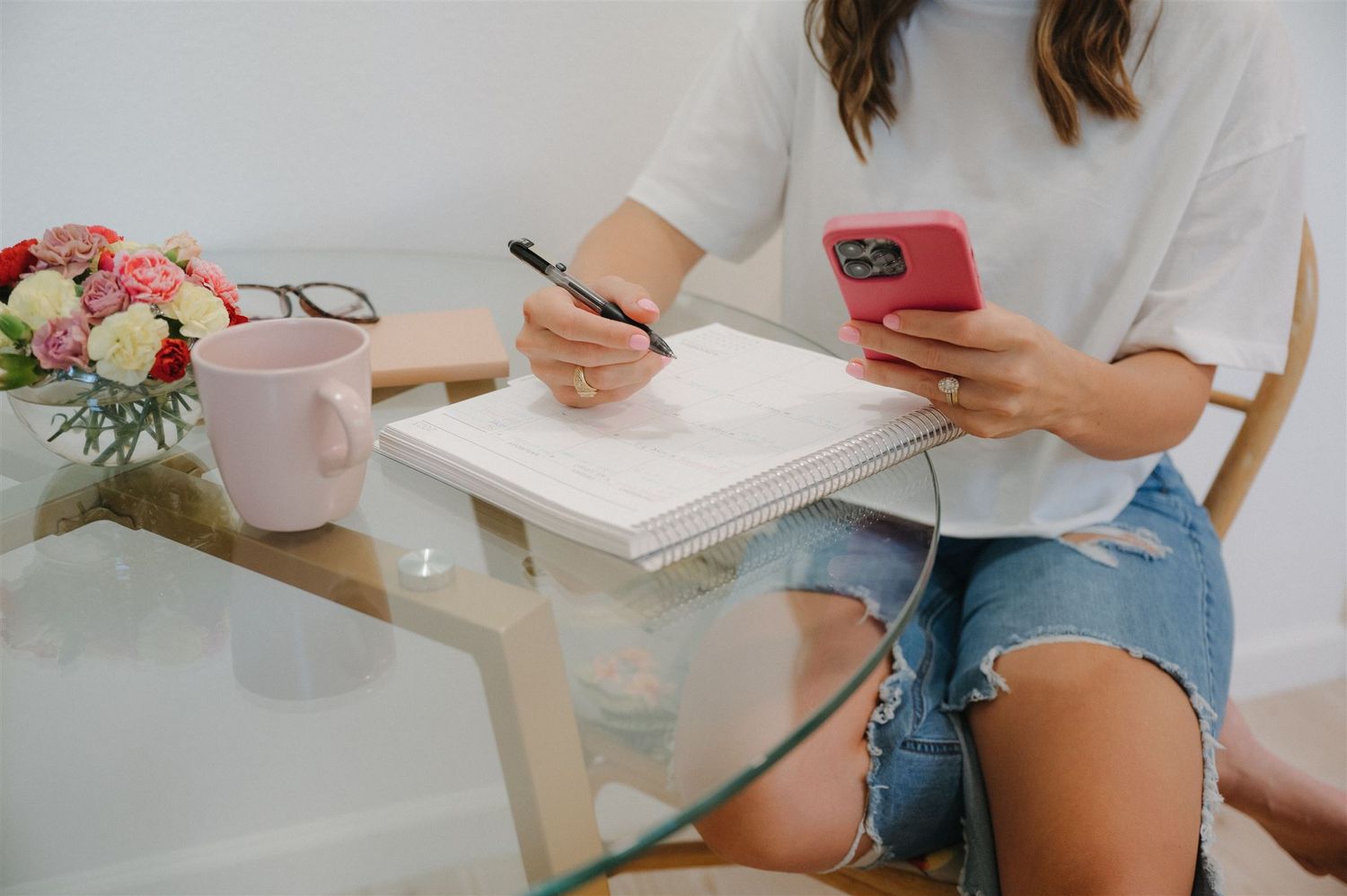 Writing in planner with phone while sitting at glass table with pink coffee mug and flowers.