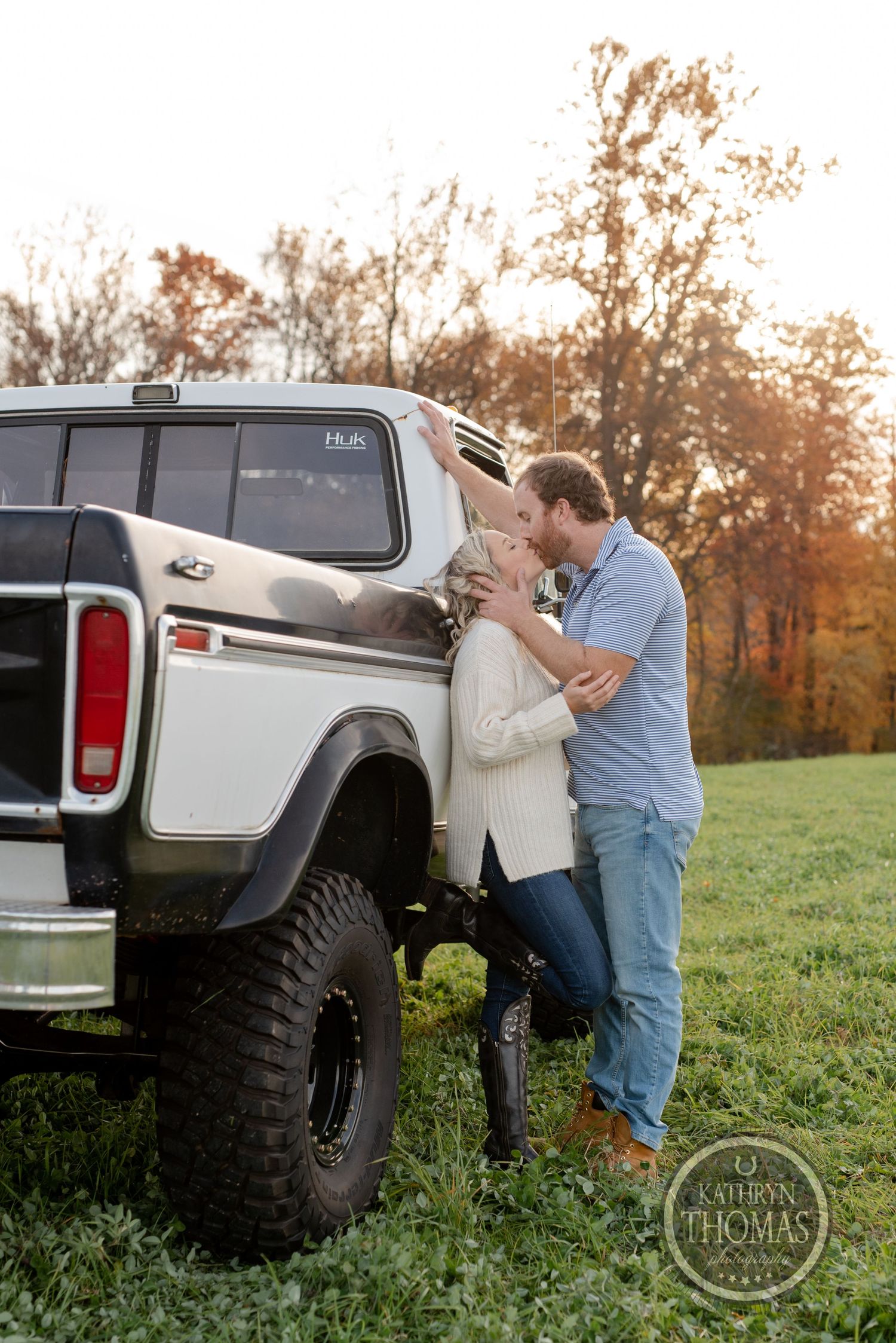 A romantic moment shared beside a lifted vintage pickup truck at sunset with fall foliage in background.