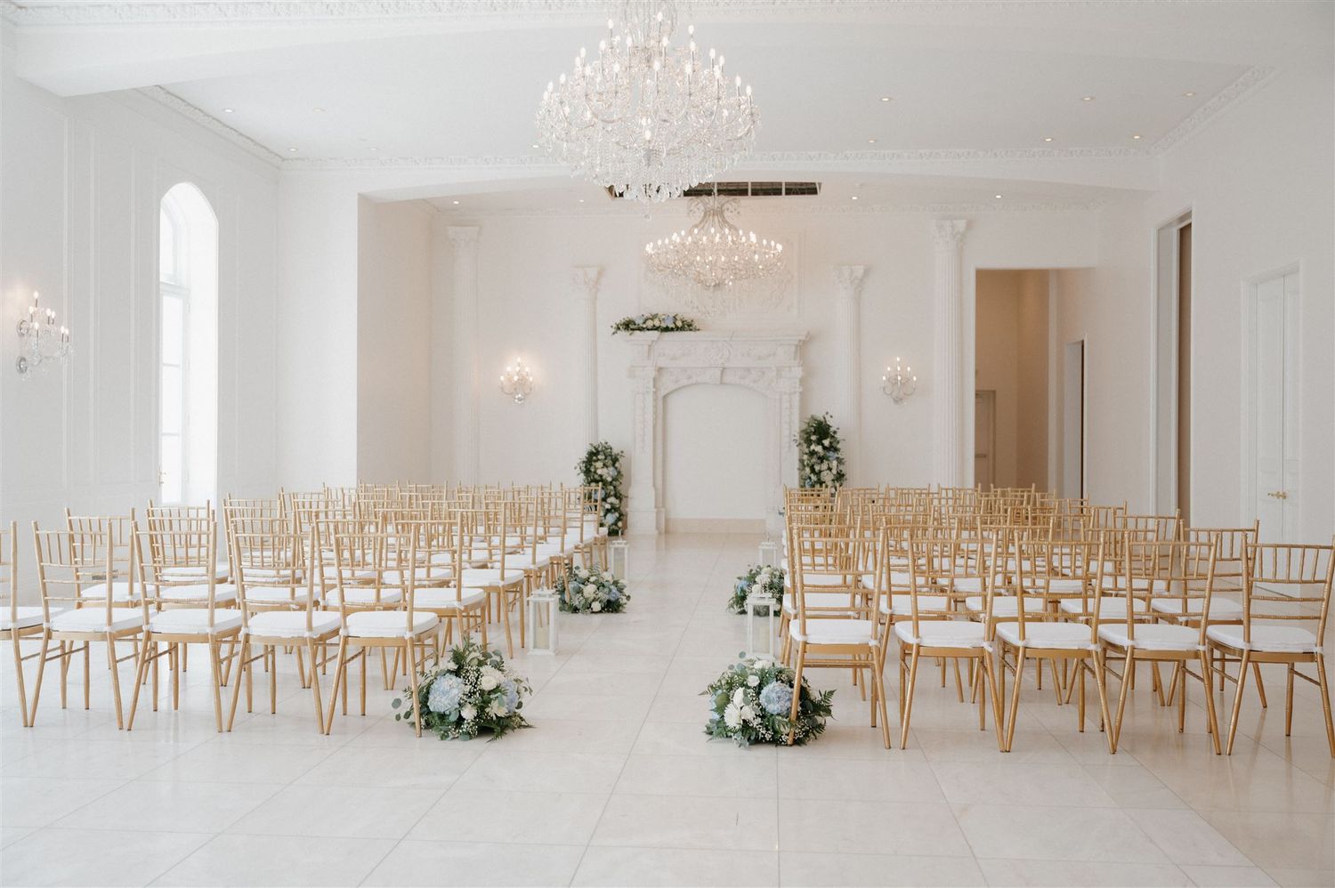 Elegant white wedding ceremony venue with gold chiavari chairs and crystal chandeliers set in rows for indoor celebration.