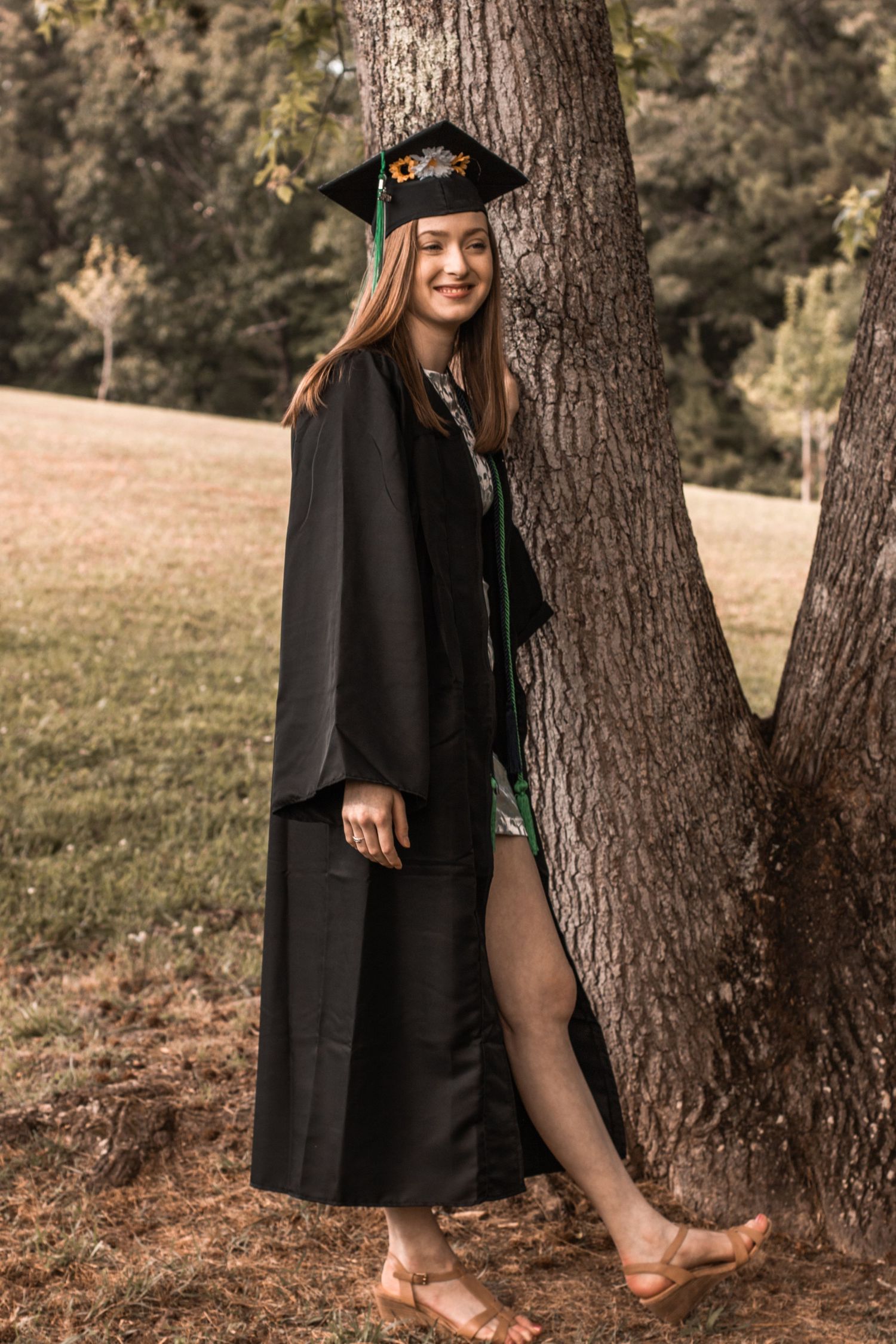 Barefoot graduate in black academic regalia leans against tree trunk in natural outdoor setting.