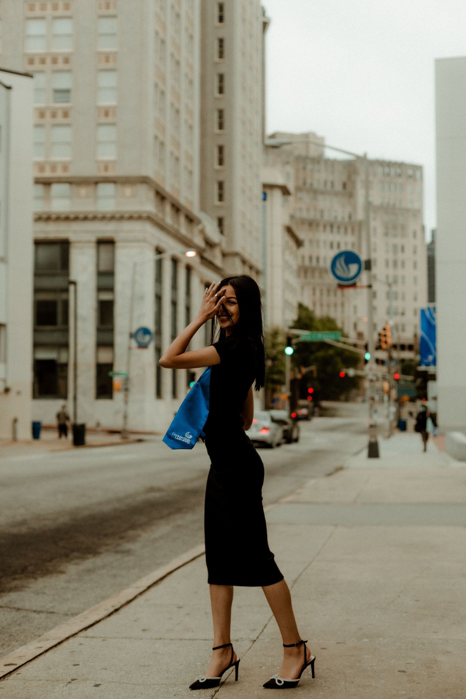 A person in a black dress poses on an urban street with tall buildings in the background.