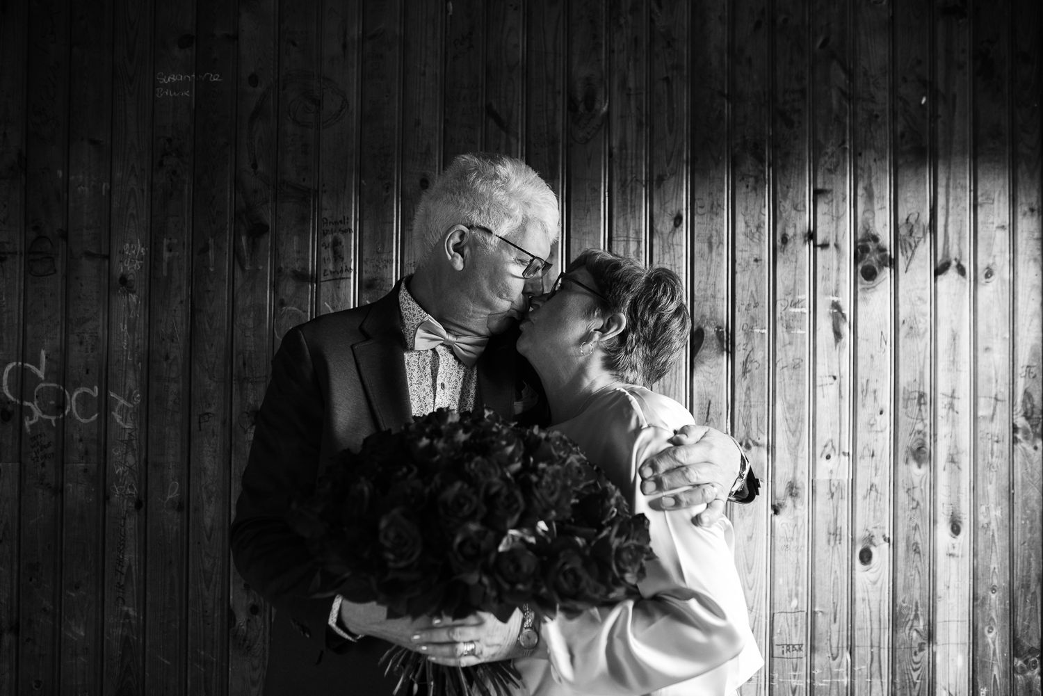 Black and white portrait of couple embracing with bouquet against wooden wall.