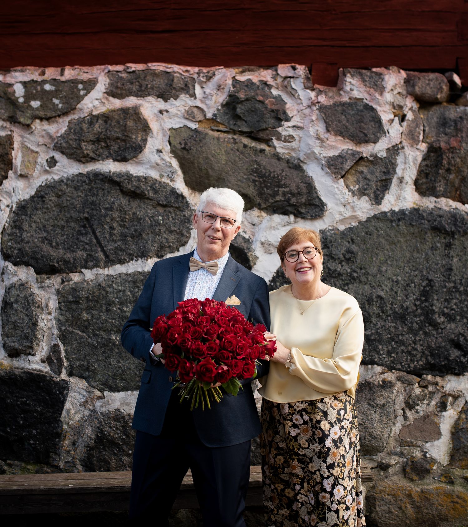 Couple posing with red roses against stone wall of historic building.