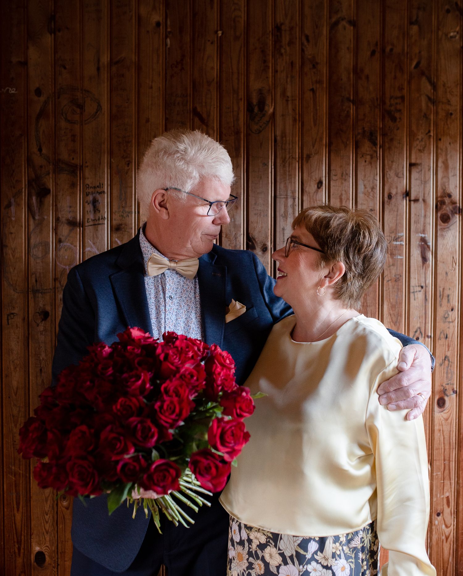 Couple sharing intimate moment with red rose bouquet against rustic wood paneling.