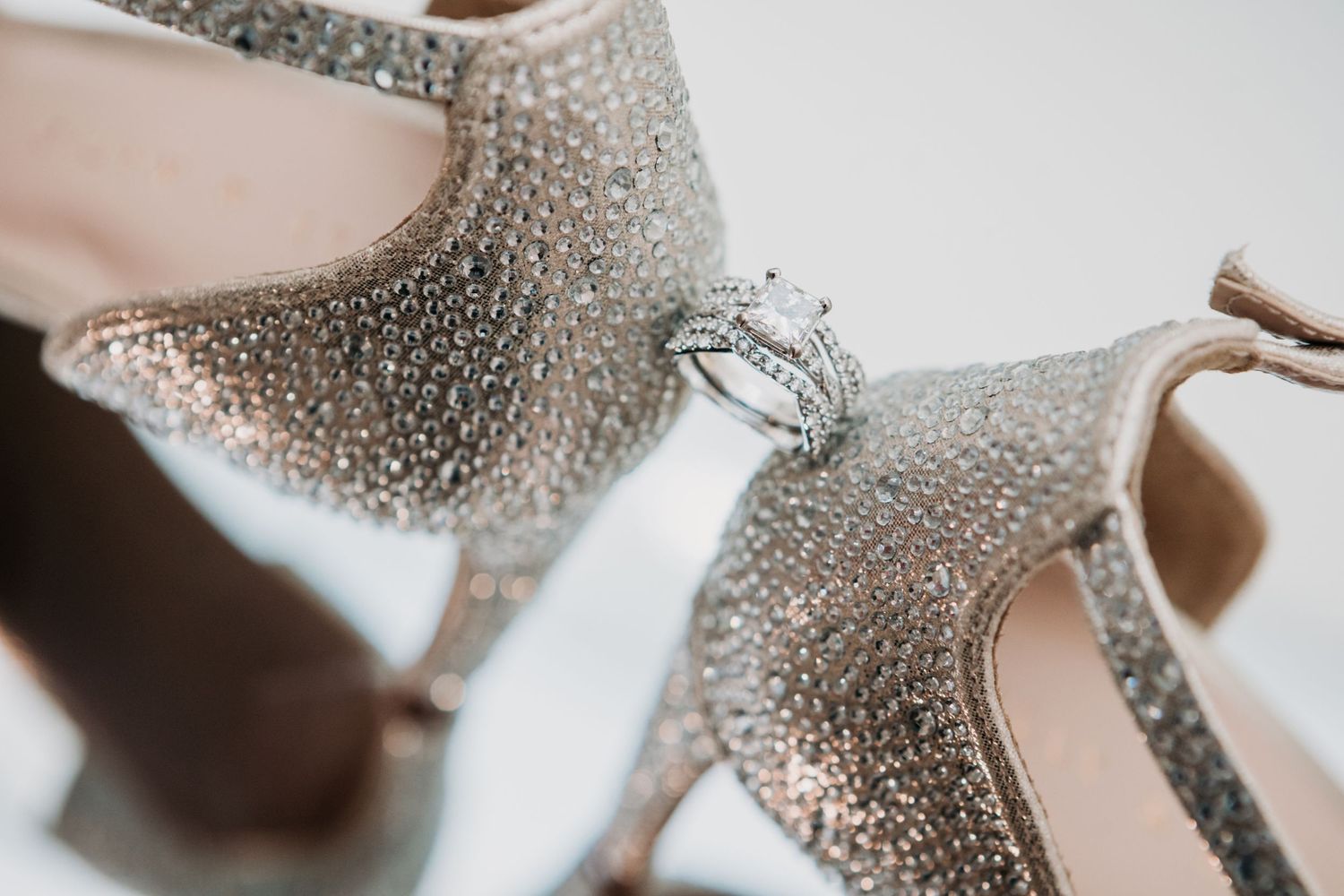 Sparkling engagement ring nestled between glittery silver wedding shoes on white background.