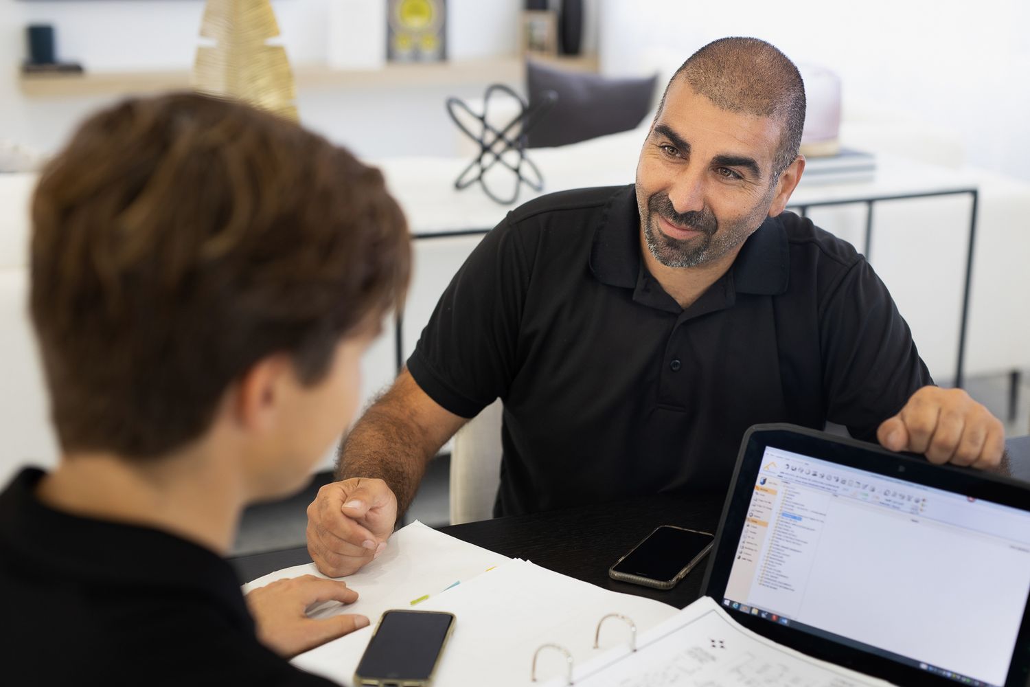 Two professionals engage in a business meeting at a modern white office desk with a laptop.
