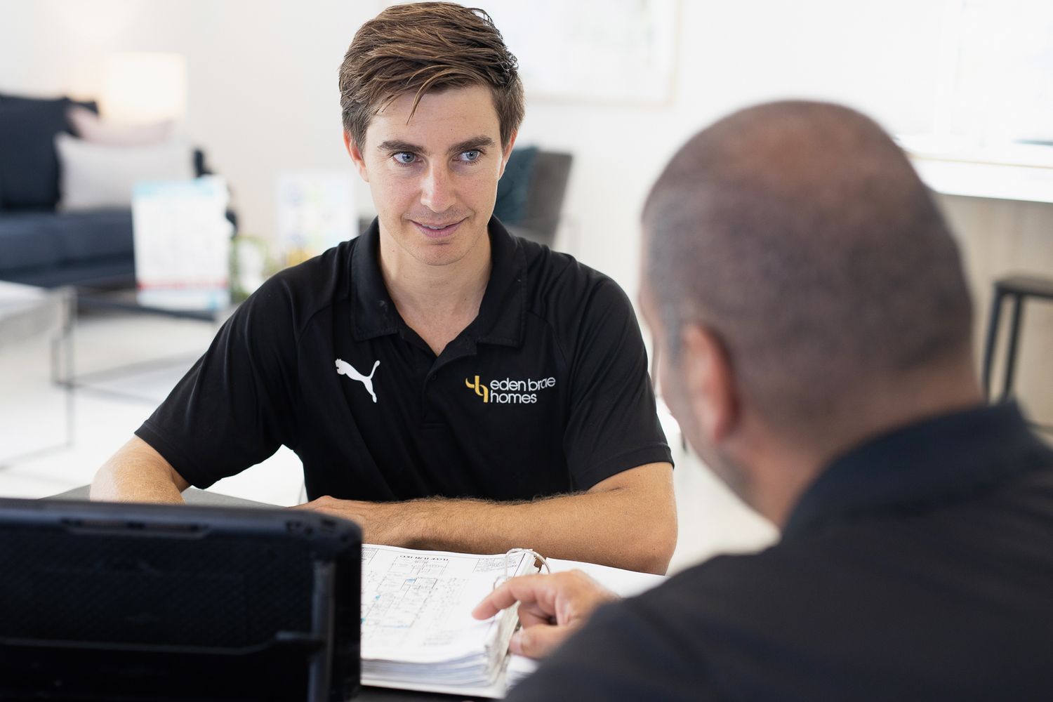A young man in a black Eden Brae Homes polo shirt consults with a client over documents at a modern desk.