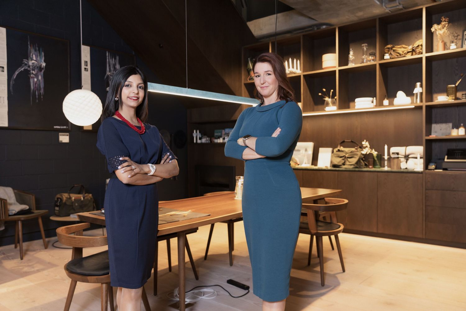 Two businesswomen in blue dresses pose with arms crossed in a contemporary office showroom.