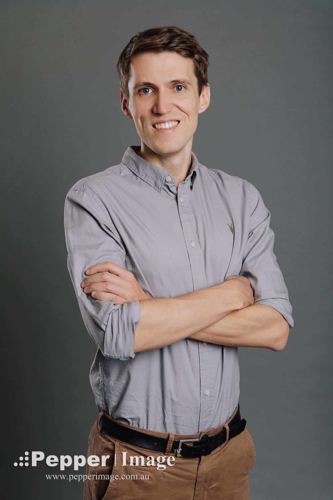 Smiling professional man in grey shirt stands with arms crossed against a grey studio background.