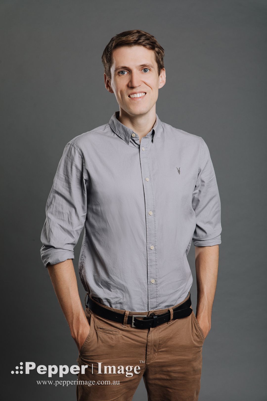 Friendly man in grey button-up shirt smiles with hands in pockets in a professional studio portrait.