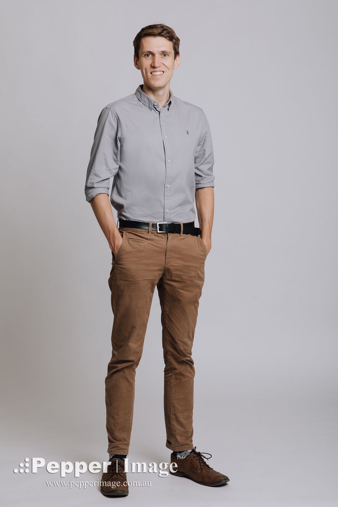 Professional man in grey shirt and brown chinos stands confidently in a clean studio setting.
