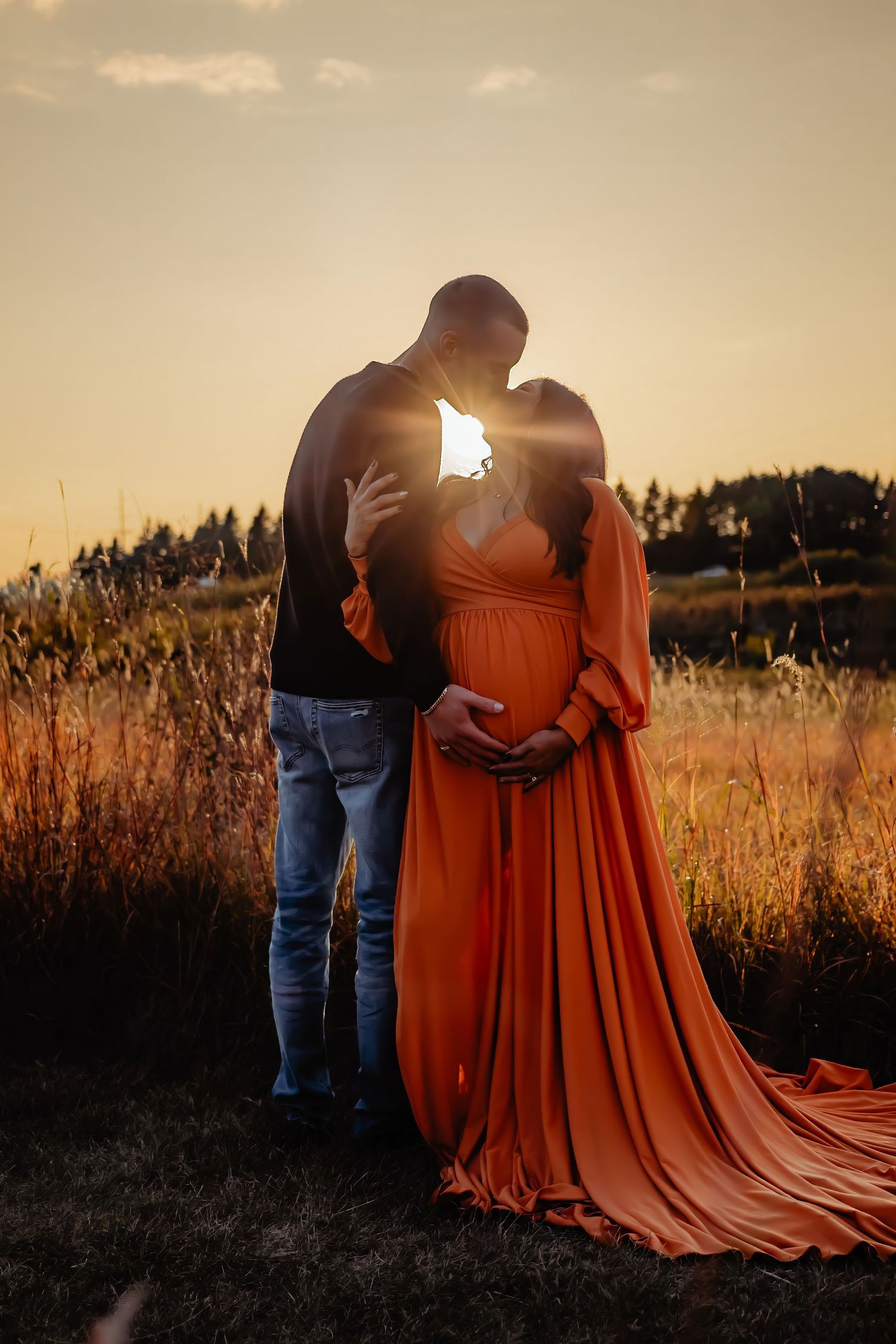 Silhouetted couple embracing at sunset in field with flowing orange maternity dress.