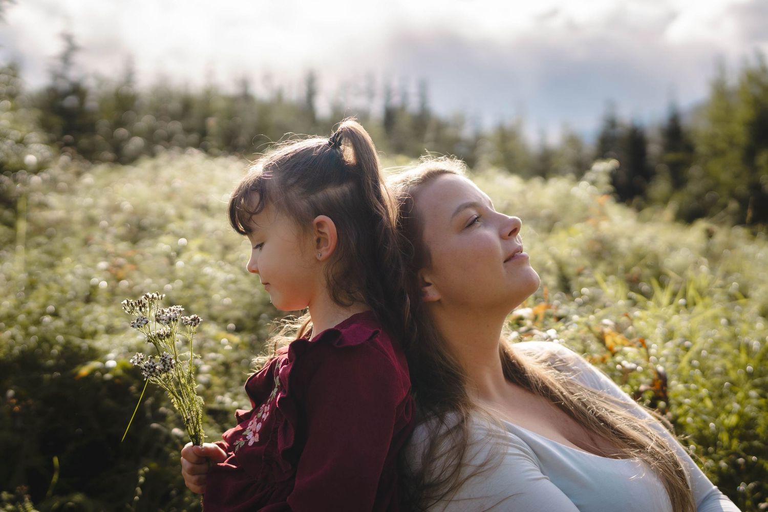 Mother and young daughter sitting back-to-back in a sunlit meadow surrounded by wildflowers and trees.