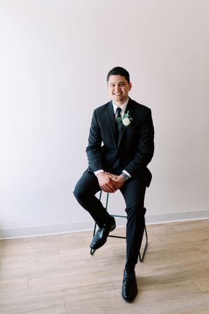 Groom portrait sitting indoors, full body, black suit. Classic and film-inspired wedding photography in Madison, Wisconsin.
