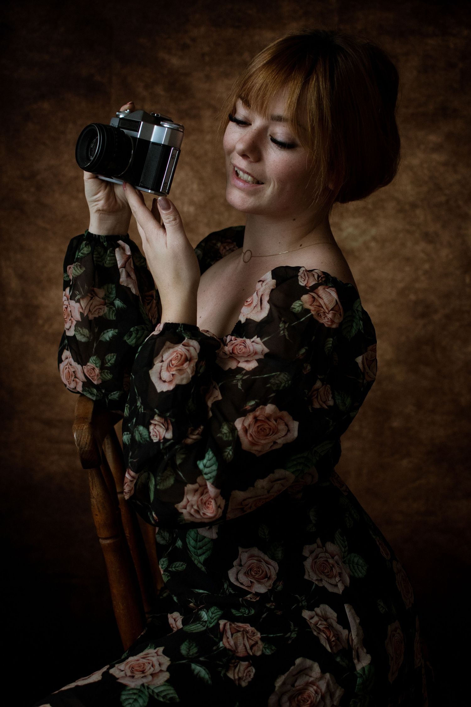 A vintage-style portrait of someone wearing a dark floral dress holding a camera against a brown background.