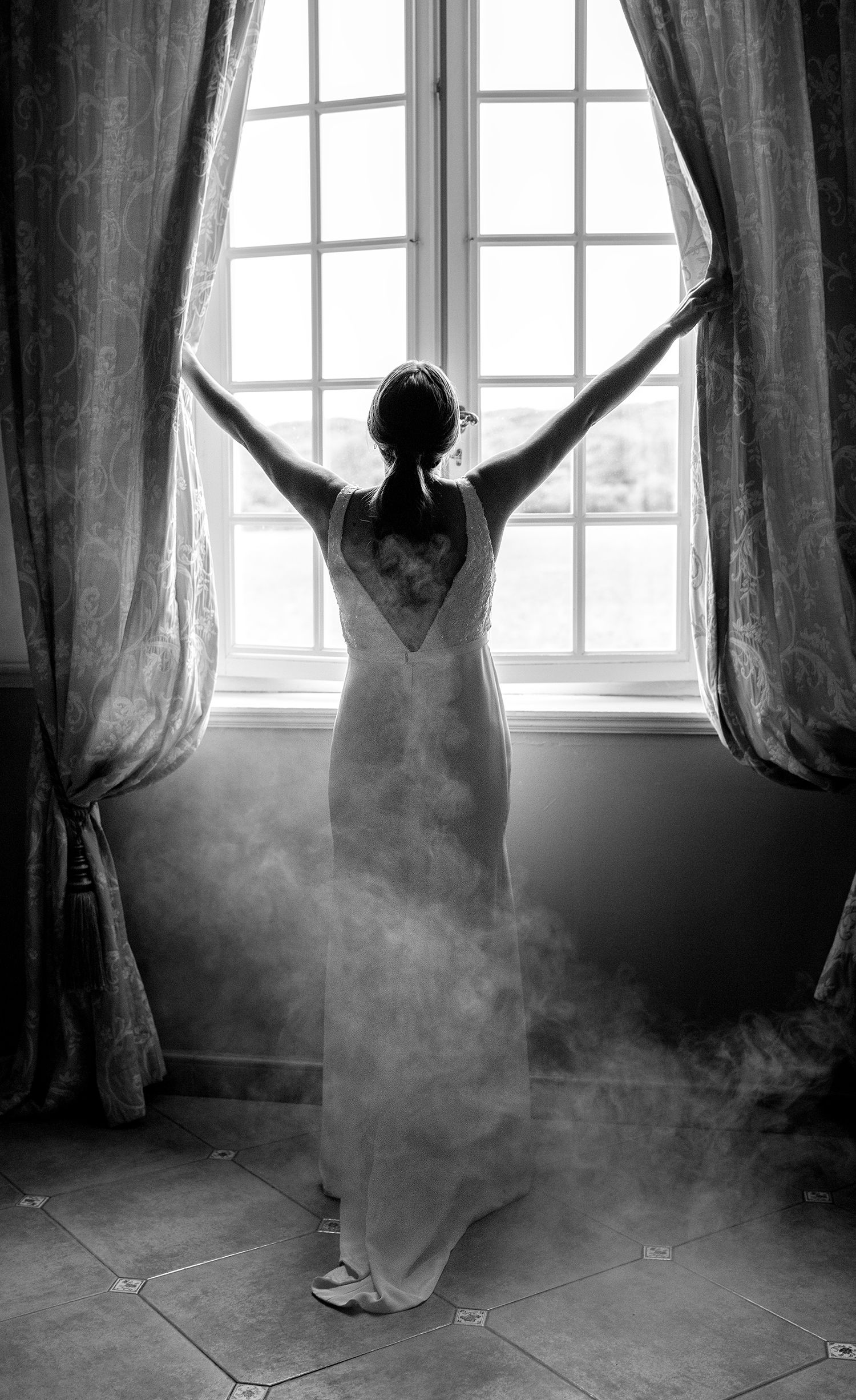 Silhouette of figure in flowing dress opening curtains with dramatic backlit window effect.